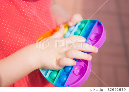Children's hands hold a silicone toy. Kid having fun with popular simple dimple multicolored rainbow colors toy. Kids being kids LGBTQ colors Vertical. 82692639