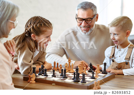Children brother and sister playing chess while sitting in living room with senior grandparents Children brother and sister playing chess while sitting in living room with senior grandparents 82693852