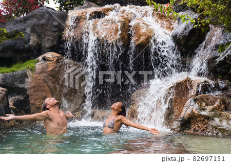 Nature couple relaxing under waterfall with open arms in freedom. People enjoying water falling in natural pool in tropical nature background. Wellness, health and spa 82697151