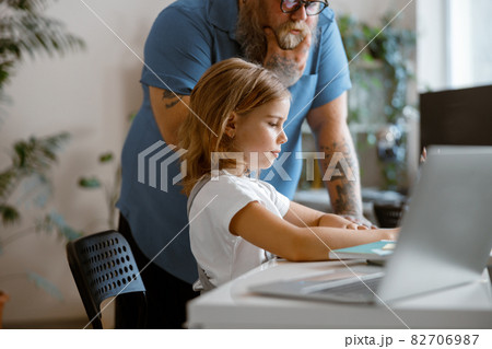 Positive little girl does homework while thoughtful daddy stands nearby in light room Positive little girl does homework while thoughtful daddy stands nearby in light room 82706987