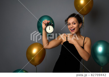 Happy woman in evening dress, rejoices at party, shows the time in the alarm clock in her hand, it's midnight. Celebrating Christmas, New Year, birthday concept on gray background with copy space Happy woman in evening dress, rejoices at party, shows the time in the alarm clock in her hand, it's midnight. Celebrating Christmas, New Year, birthday concept on gray background with copy space 82709442