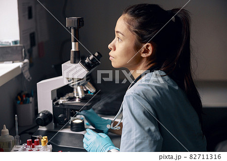 Thoughtful Asian lab assistant sits near microscope looking out of window at workplace 82711316