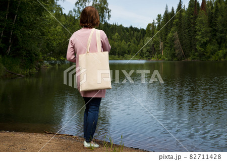 Woman standing on the river bank in pink jacket with tote bag mockup . Woman standing on the river bank in pink jacket with tote bag mockup . 82711428