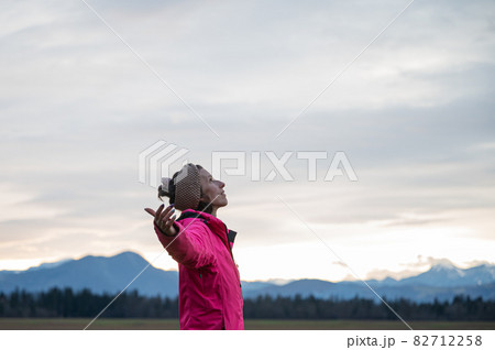 Young woman in pink jacket standing under evening sky with her arms spread widely 82712258