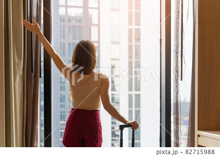 Portrait rear view of woman standing close to the window, looking outside in hotel room after check-in 82715988