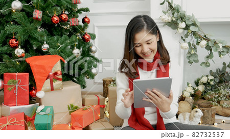 Asian woman using smart tablet sit at christmas tree xmas present gift box in cozy living room. Woman hand touch screen smile shopping online, happiness time christmas holiday winter season festive 82732537