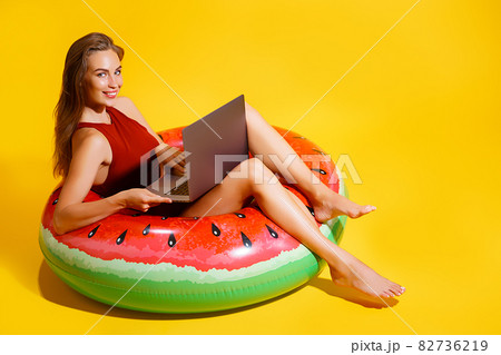 Smiling girl in swimsuit sits in swim inflatable ring while holds laptop pc. Summer vacation rest 82736219