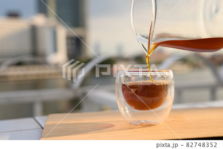 Close up pouring hot black coffee from the coffee pot into a cup on the balcony by the river. Copy space. 82748352