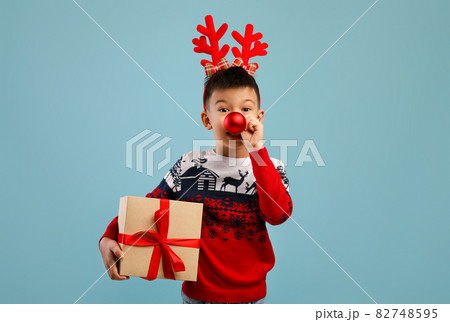 Holiday Fun. Asian Boy Wearing Deer Horns And Christmas Sweater Holding Present 82748595