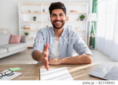 Employment concept. Arab businessman stretching hand for handshake, greeting to camera, sitting at workplace at home 82748681