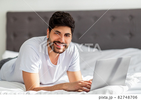 Always online. Young arab man working on laptop distantly, lying on bed and smiling to camera, free space Always online. Young arab man working on laptop distantly, lying on bed and smiling to camera, free space 82748706
