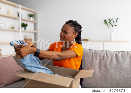 Dissatisfied black female customer opening box from online store, unhappy with received jeans at home 82748754