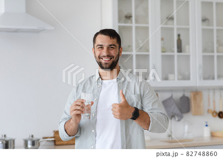 Smiling adult european man with beard holding glass of water and showing thumb up at kitchen interior 82748860