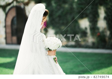 Beautiful bride in elegant white dress holding bouquet posing in park 82749074
