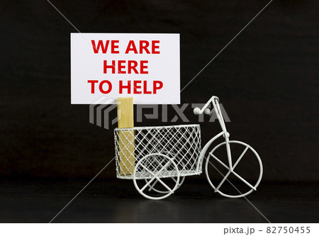 Support symbol. Wooden clothespin with white sheet of paper. Words 'we are here to help'. Miniature bicycle model. Beautiful black background. Business, support concept, copy space. Support symbol. Wooden clothespin with white sheet of paper. Words 'we are here to help'. Miniature bicycle model. Beautiful black background. Business, support concept, copy space. 82750455