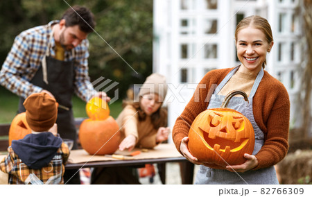 Smiling woman with jack-o-lantern smiling at camera while carving pumpkins with family in backyard 82766309