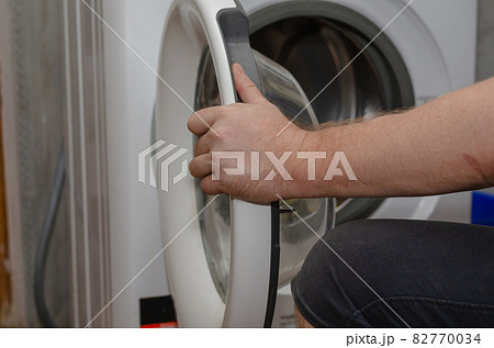 An adult man next to a washing machine. A hand closes the door o An adult man next to a washing machine. A hand closes the door o 82770034
