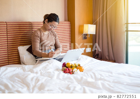 Young Asian women smiling happily at freelance work, working on a notebook while relaxing in bed with snacks and fruit in a hotel room. Vacation and relaxation Young Asian women smiling happily at freelance work, working on a notebook while relaxing in bed with snacks and fruit in a hotel room. Vacation and relaxation 82772551