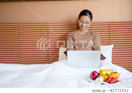 Young Asian women smiling happily at freelance work, working on a notebook while relaxing in bed with snacks and fruit in a hotel room. Vacation and relaxation 82772552