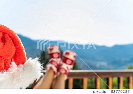 Woman in socks, boots and Christmas red hat sitting on a balcony in the mountains putting her feet on the wooden terrace railing 82776287