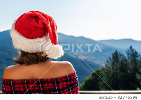 Woman in Christmas red hat wrapped in a blanket standing on a balcony in the mountains 82776598
