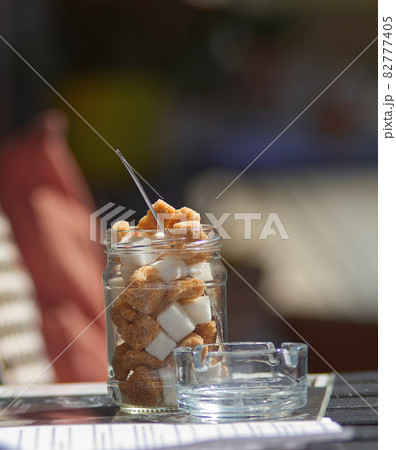 A jar of cube sugar, brown and white cube sugar in a jar on a coffee table in the street, with a small ashtray next to it. Blurred background. 82777405