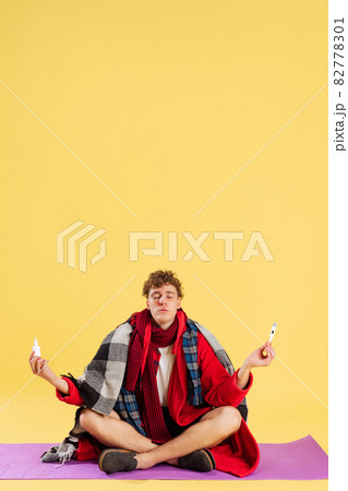 Cropped portrait of young sick man wearing plaid, robe and slippers sitting on meditation pose isolated over yellow background Cropped portrait of young sick man wearing plaid, robe and slippers sitting on meditation pose isolated over yellow background 82778301