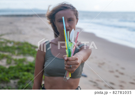 Girl stands on the beach near the ocean and holds used plastic dishes in one hand. Clean ocean beach on the background. Single-use plastic waste Girl stands on the beach near the ocean and holds used plastic dishes in one hand. Clean ocean beach on the background. Single-use plastic waste 82781035