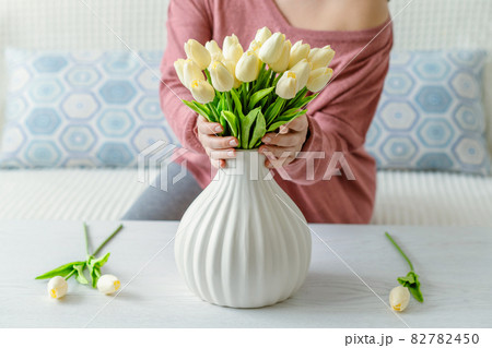 Woman putting white tulips flowers in vase sitting at the living room coffee table. Lifestyle Woman putting white tulips flowers in vase sitting at the living room coffee table. Lifestyle 82782450