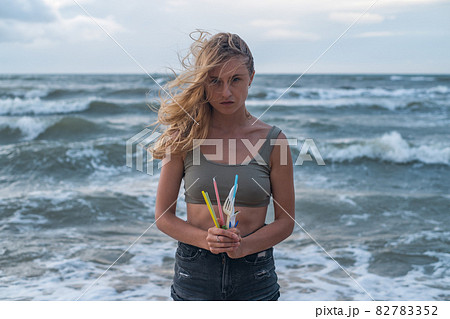 Portrait of Sad blond caucasian young woman standing on the beach and holding used plastic dishes and straws. Sea on the background. plastic pollution Portrait of Sad blond caucasian young woman standing on the beach and holding used plastic dishes and straws. Sea on the background. plastic pollution 82783352