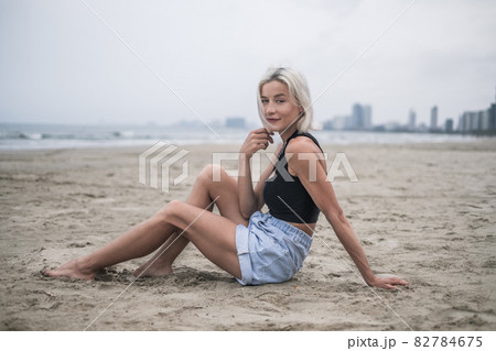 Beautiful fit caucasian young woman with blond hair, wearing denim shorts, short top and stylish necklace. Sitting on the beach. Model posing 82784675