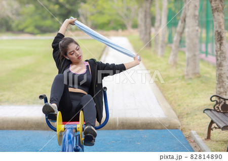 Happy smiling young beautiful asian woman in sportswear relaxing on public exercise machine. Cute and sporty girl working out on bike during morning in summer outdoor. 82814890