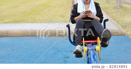 Happy smiling young beautiful asian woman in sportswear relaxing on public exercise machine. Cute and sporty girl working out on bike and using mobile smartphone outdoor 82814891
