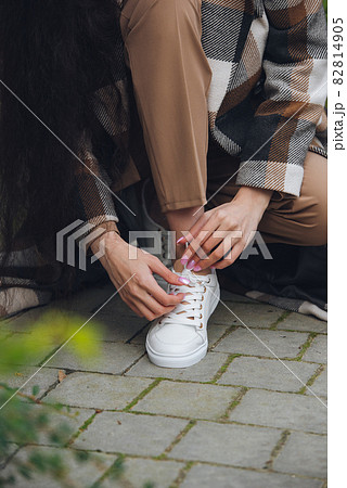 Closeup photo of young woman wearing checkered long coat, and beige pants . Lady posing on city street. Women ties laces on sneakers Closeup photo of young woman wearing checkered long coat, and beige pants . Lady posing on city street. Women ties laces on sneakers 82814905