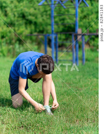 teenage boy exercising outdoors, sports ground in the yard, he ties his shoelaces and does a warm-up, healthy lifestyle teenage boy exercising outdoors, sports ground in the yard, he ties his shoelaces and does a warm-up, healthy lifestyle 82841163