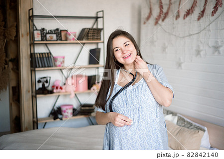 A pregnant girl listens with a phonendoscope to her baby in her belly and smiles. 82842140