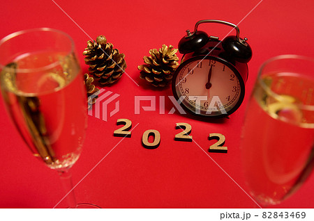 Composition with wooden numerals, 2022 year, black alarm clock with midnight on the clock face, golden pine cones and champagne flutes with a sparkling wine on a red background with copy space for ad 82843469