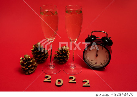 Christmas and New Year concept. Champagne flutes with a sparkling wine, alarm clock showing midnight in the face clock, golden pine cones and wooden numerals 2022, on red background with copy space 82843470