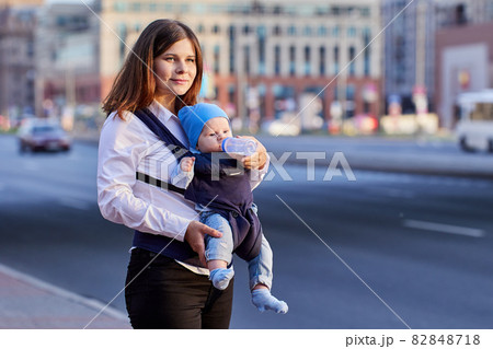 Woman is feeding baby in sling from bottle outdoors. 82848718