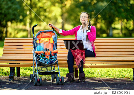 Teleworking by woman with infant in baby carriage with help of laptop outdoors. 82850955