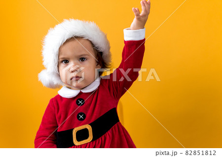 Serious Portraite Cute Happy Cheerful Chubby Baby Girl in Santa Hat Waving Hand Up At Yellow Background. Child Play Christmas Scene Celebrating Birthday. Kid Have Fun Spend New Year Time Copy Space 82851812