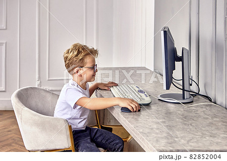 Happy european little boy in glasses sits behind computer. Happy european little boy in glasses sits behind computer. 82852004