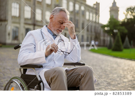 Tired senior handicapped male doctor in wheelchair wearing lab coat taking off his glasses, having headache, posing outdoors Tired senior handicapped male doctor in wheelchair wearing lab coat taking off his glasses, having headache, posing outdoors 82853389