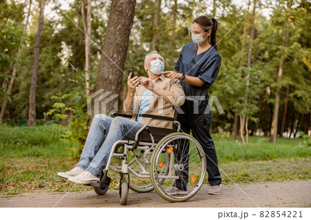 Young nurse in face shield and protective face mask assisting senior handicapped man in wheelchair using his smartphone during a walk in the park Young nurse in face shield and protective face mask assisting senior handicapped man in wheelchair using his smartphone during a walk in the park 82854221
