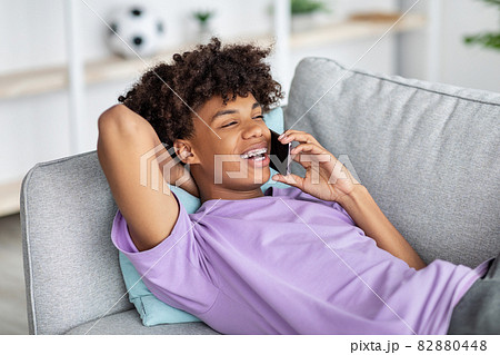 Cheerful black teenager lying on couch, having smartphone conversation with friend at home 82880448