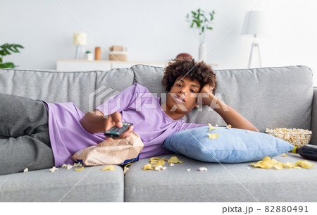 Lazy black teen guy lying on sofa with scattered food, holding TV controller, watching dull movie on television at home Lazy black teen guy lying on sofa with scattered food, holding TV controller, watching dull movie on television at home 82880491