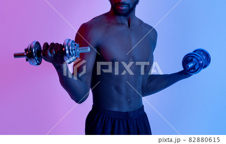 Bodybuilding and strength workout. Unrecognizable black guy with bare torso training with dumbbells in neon light Bodybuilding and strength workout. Unrecognizable black guy with bare torso training with dumbbells in neon light 82880615