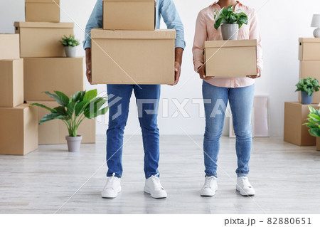 Young african american husband and wife carry cardboard boxes, potted plants in living room 82880651