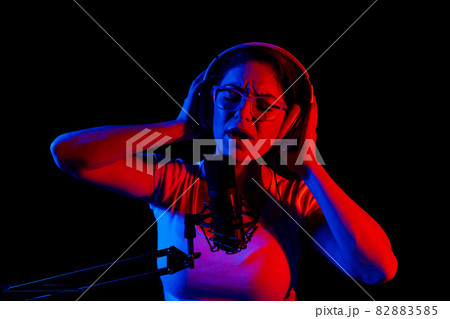 Caucasian woman in glasses and headphones sings into a microphone in neon light on a black background. An emotional girl is recording a song in a recording studio 82883585