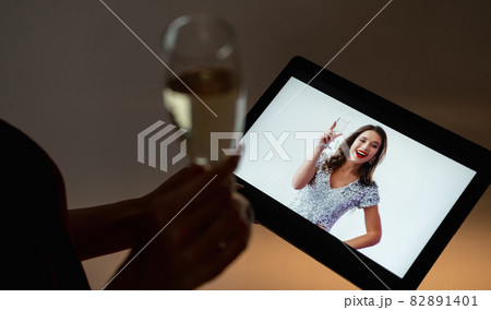 Online birthday. Festive greeting. Virtual party. Unrecognizable woman with glass of champagne looking happy female friend congratulating on tablet computer in dark room interior. 82891401
